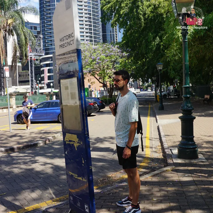 Reading a 'Historic Precinct' information panel on a central Brisbane street, with jacarandas in bloom
