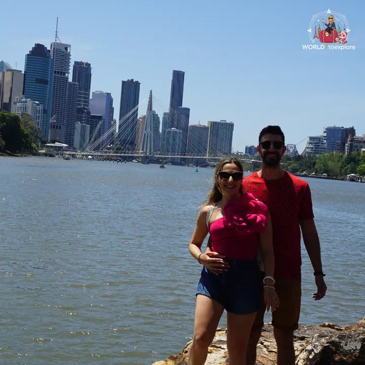 A couple on the rocks by the river, with the Brisbane skyline and the Neville Bonner Bridge in the background