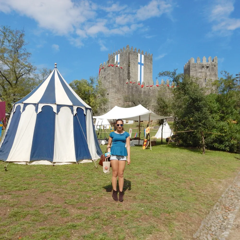 Woman in a blue top standing by a blue-and-white striped medieval tent, with the castle keep decked out in red banners behind her