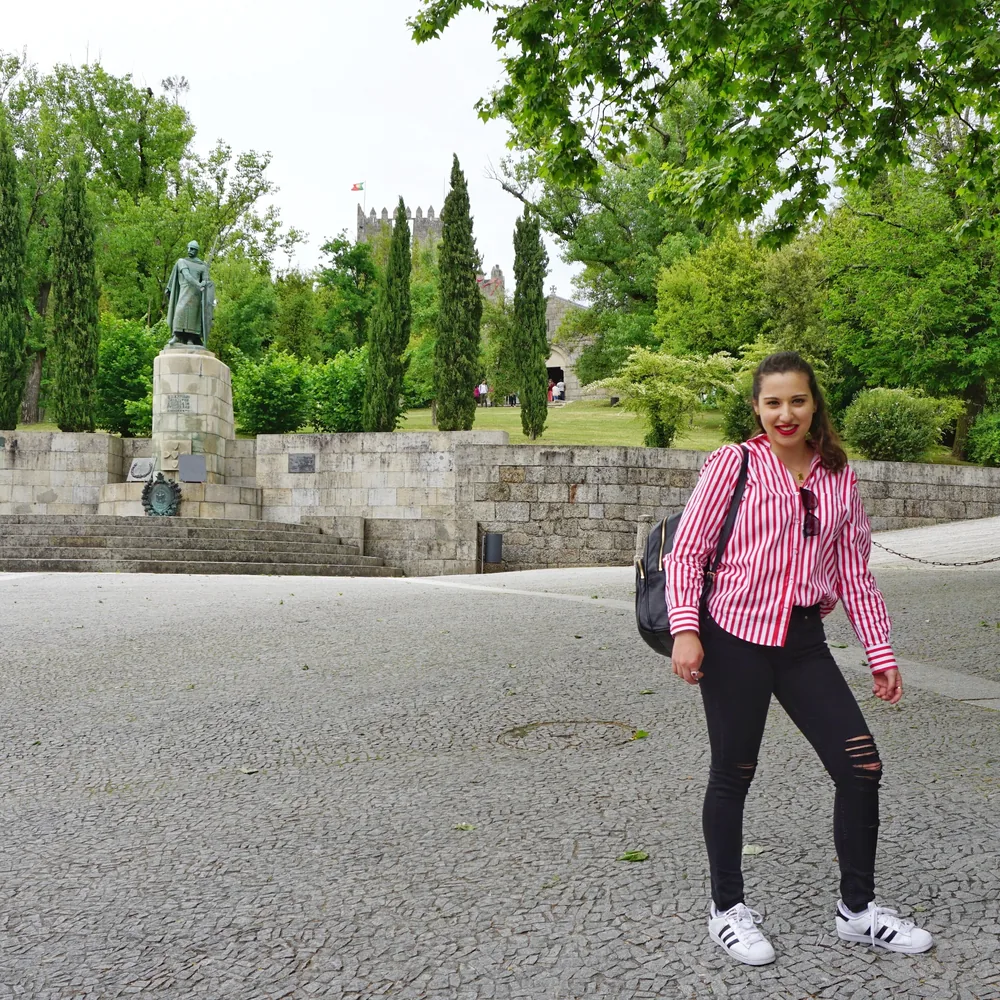 Woman in a red-and-white striped shirt and ripped black jeans standing by the D. Afonso Henriques statue, with the castle tower in the background