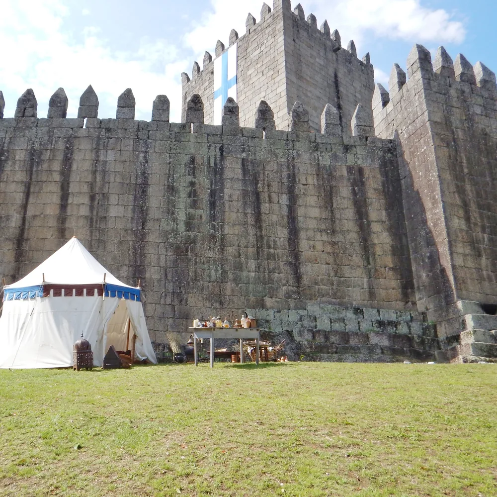 The castle wall seen from below, with a blue cross banner hanging from the battlements and a medieval tent set up on the lawn in front