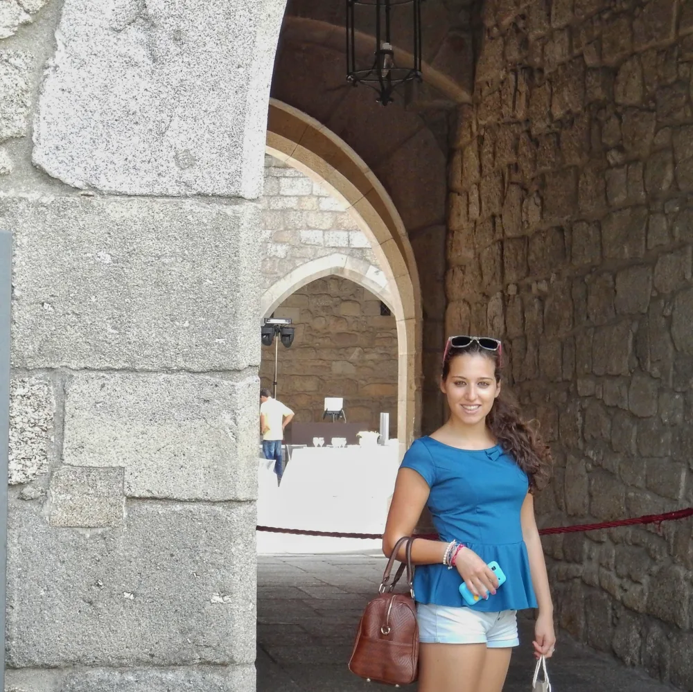Woman in a blue top smiling under the castle's stone entrance archway, holding a brown handbag and her phone