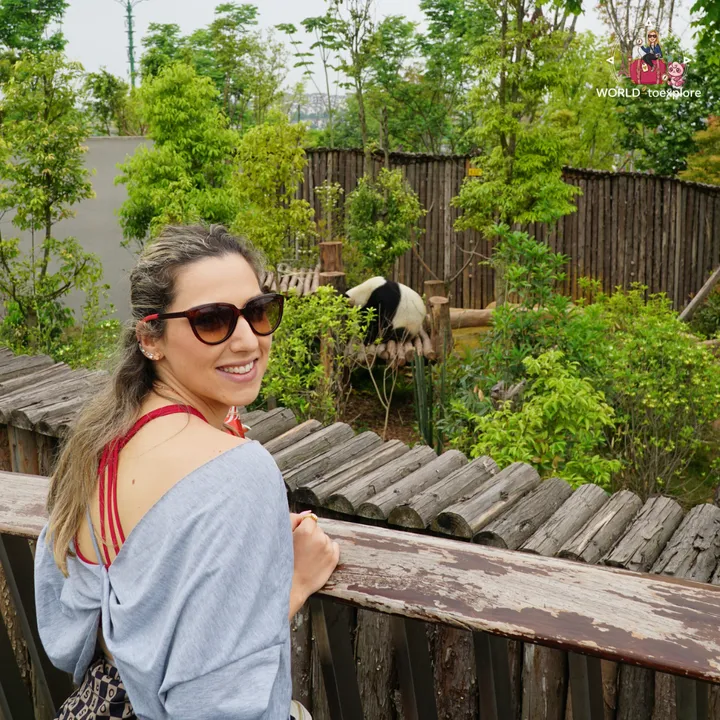 Cristina smiling next to a wooden fence, with a giant panda walking over logs in the background