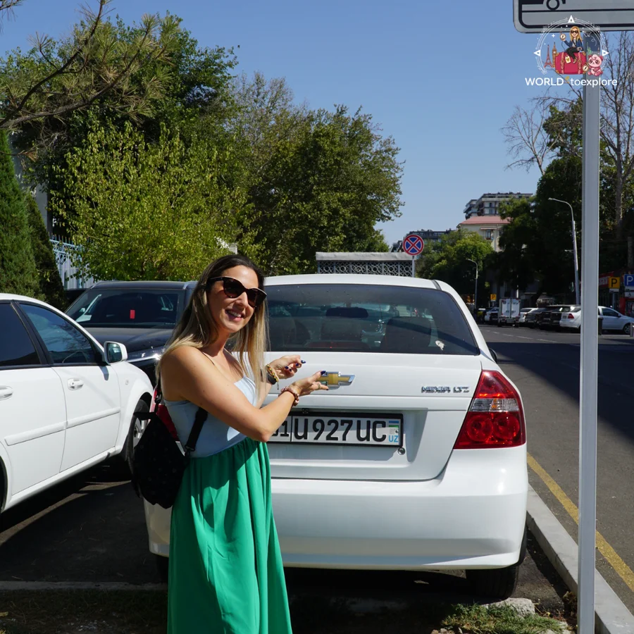 Woman in a green skirt and light blue top, smiling, pointing at the badge on the back of a white Chevrolet with an Uzbek number plate