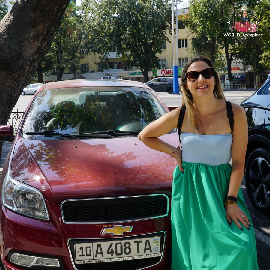 Woman in sunglasses with a hand on her hip in front of a red Chevrolet with an Uzbek number plate, on a tree-lined street