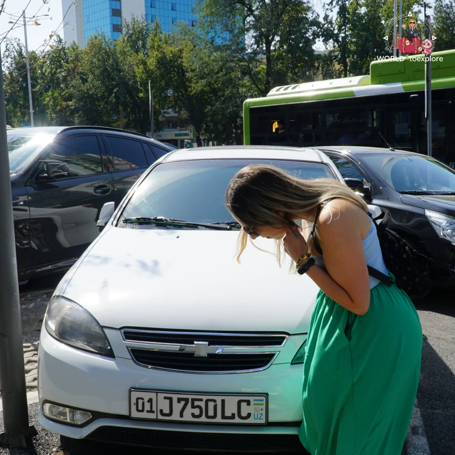 Woman bent over laughing next to the front of a white Chevrolet, with dark cars and a green bus behind her