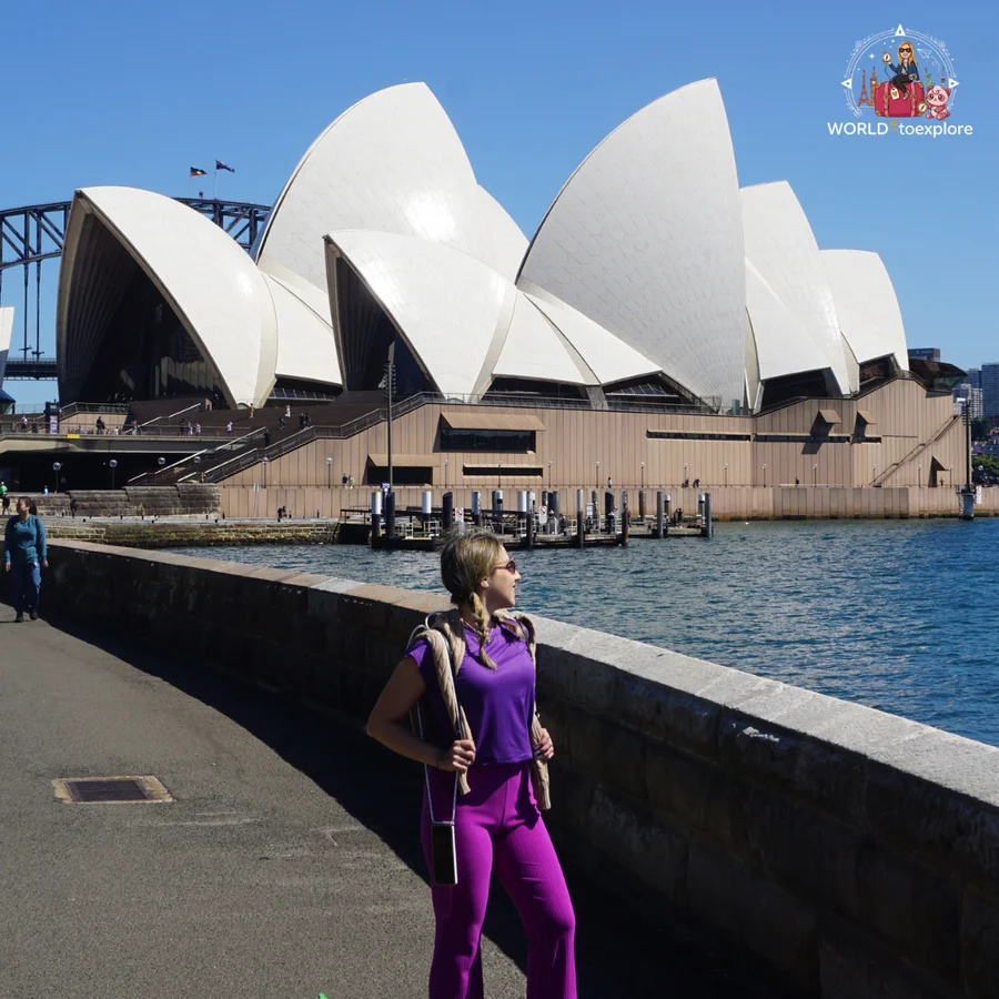 By the stone wall along the Sydney waterfront, with the white sails of the Opera House and the Harbour Bridge behind on a clear blue day