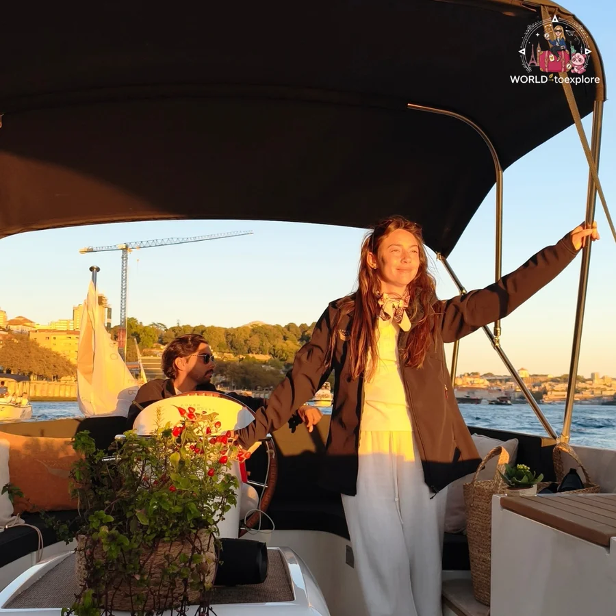Inês, in a brown jacket and white clothes, leaning on the Wondy's canopy, with a man at the helm behind her and the golden light of the late afternoon