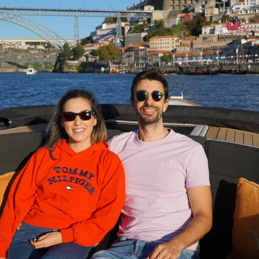 Cristina and her husband sitting side by side aboard the Wondy, smiling, with the Ribeira in Porto and the Luís I Bridge behind them