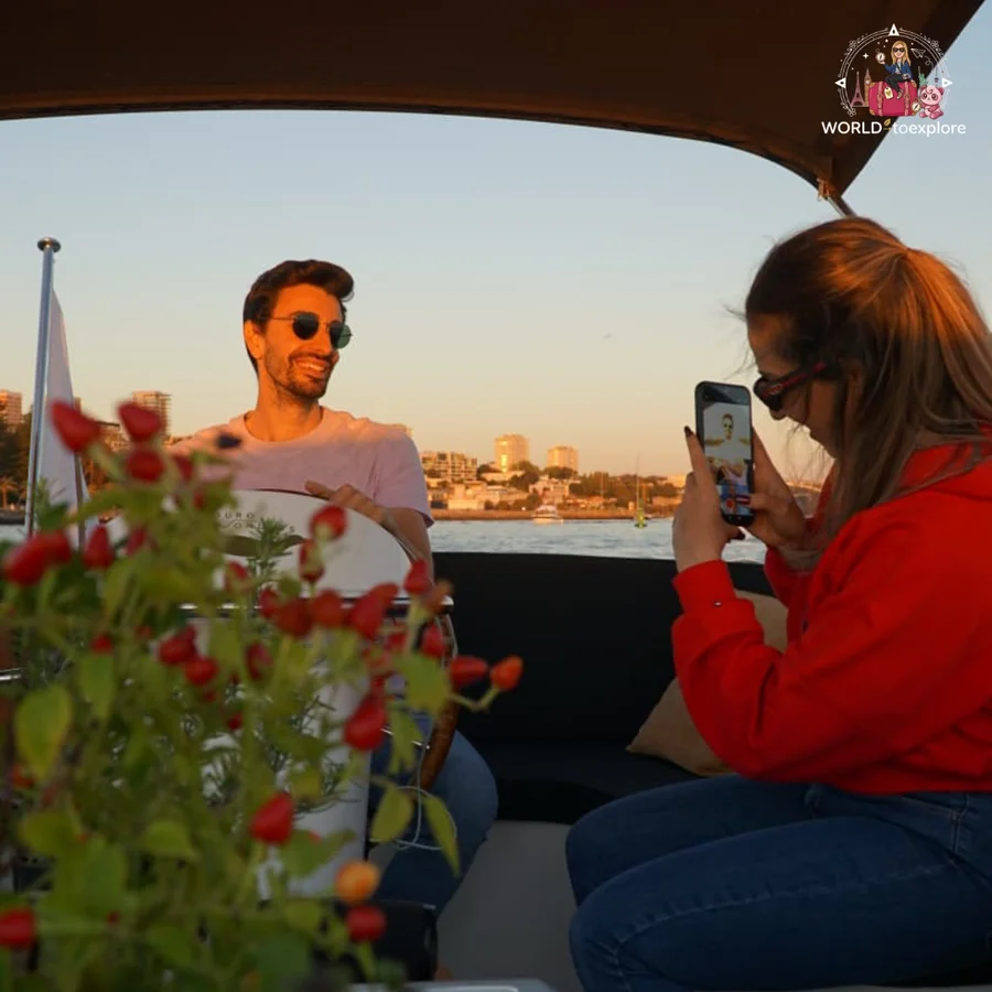 Her husband at the helm of the Wondy smiling while Cristina takes a photo with her phone, among the red berries of a plant at sunset