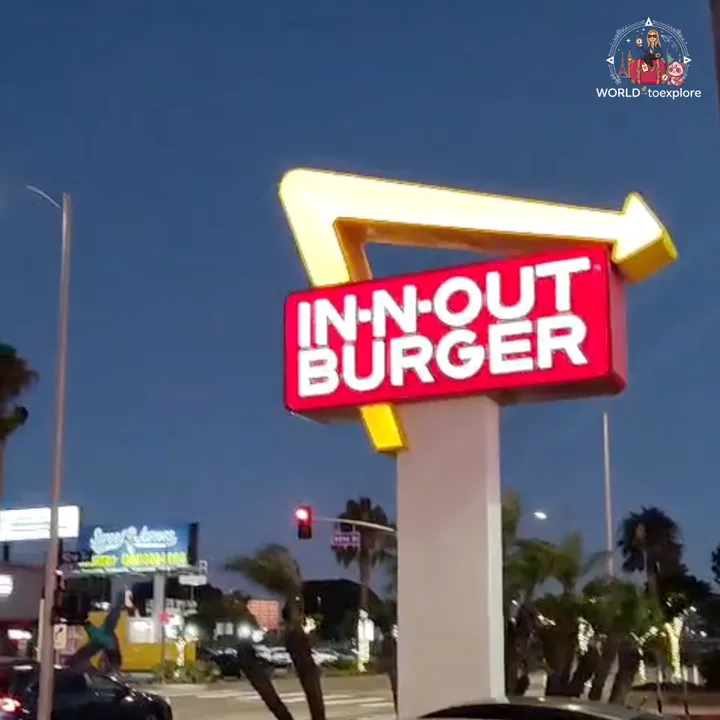 The illuminated In-N-Out Burger sign, shaped like a yellow arrow, lit up at dusk next to the road