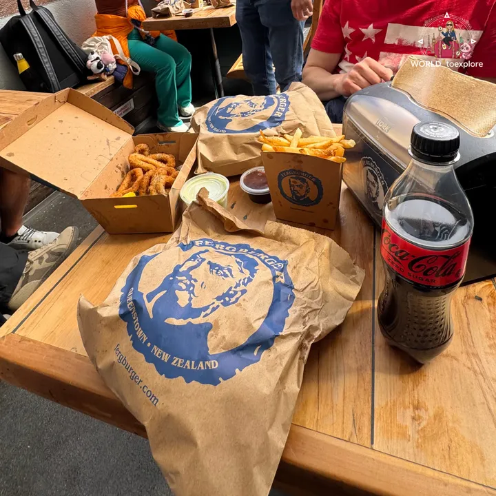 A Fergburger paper bag with the restaurant's logo, next to fries, breaded onion rings and a bottle of Coca-Cola Zero on an outdoor wooden table