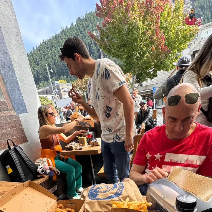 A group sitting on an outdoor terrace in Queenstown eating Fergburger burgers, with pine-covered mountains and autumn trees in the background