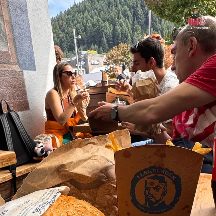 A group at a table sharing fries from a box with the Fergburger logo, with Queenstown's forested mountain in the background