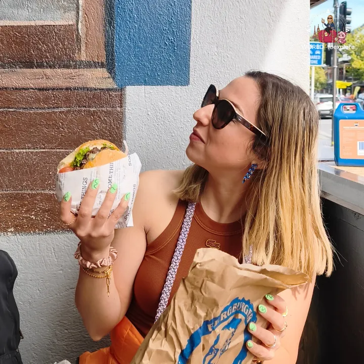The author holding a Fergburger burger wrapped in paper, with the brand's paper bag in her other hand, leaning against a wall painted blue and brown