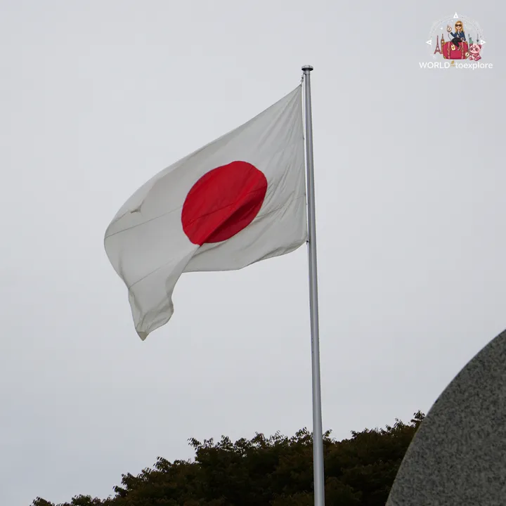The Japanese flag flying in the Peace Memorial Park, under an overcast sky, with the curved stone of the cenotaph on the right