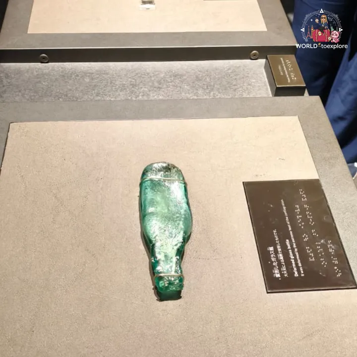 A green glass bottle deformed by the heat, on the base of the display case with its label beside it