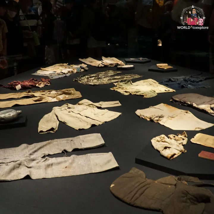A display case with victims' clothing laid out against a dark background and visitors reflected in the glass