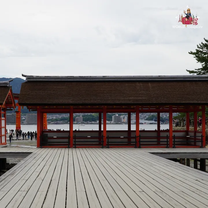 A wooden boardwalk at the shrine and a pavilion with red pillars, facing the sea and the city of Hiroshima in the distance