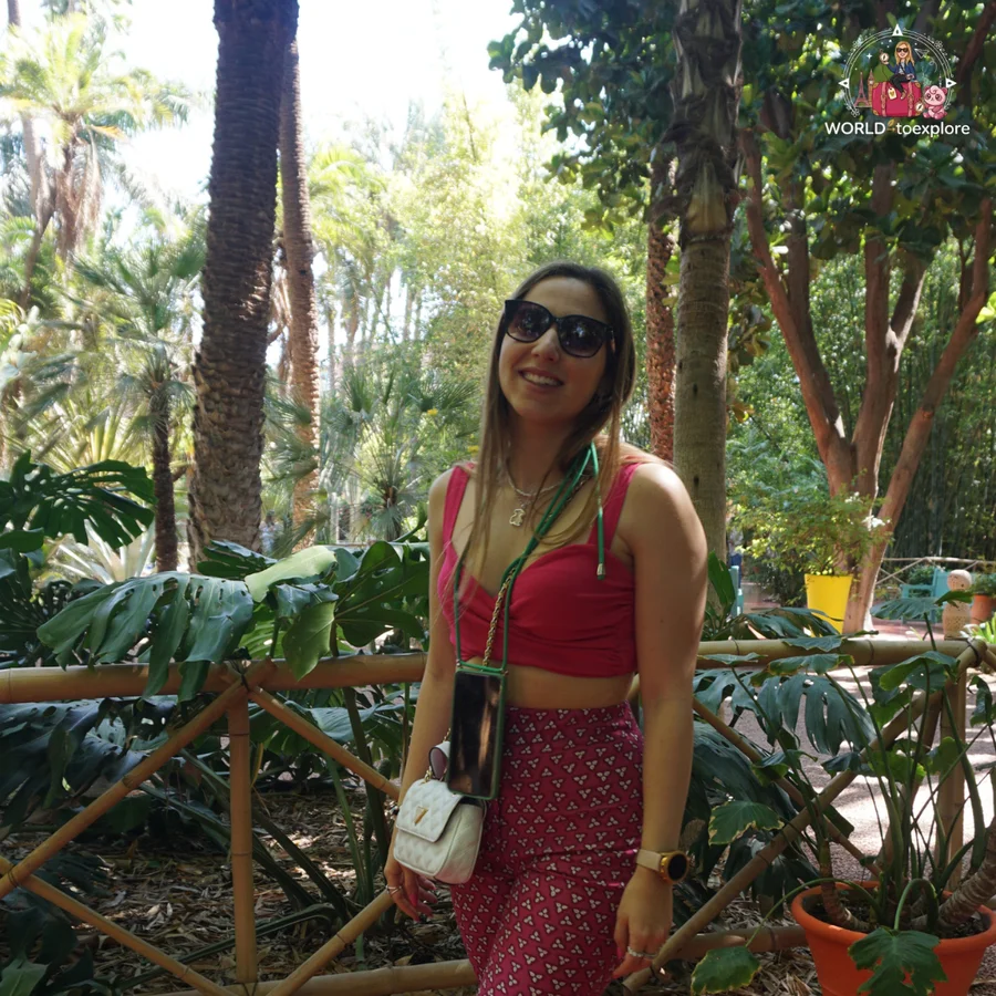 Smiling woman in sunglasses in front of a bamboo railing, with tropical foliage and a plant in an orange pot