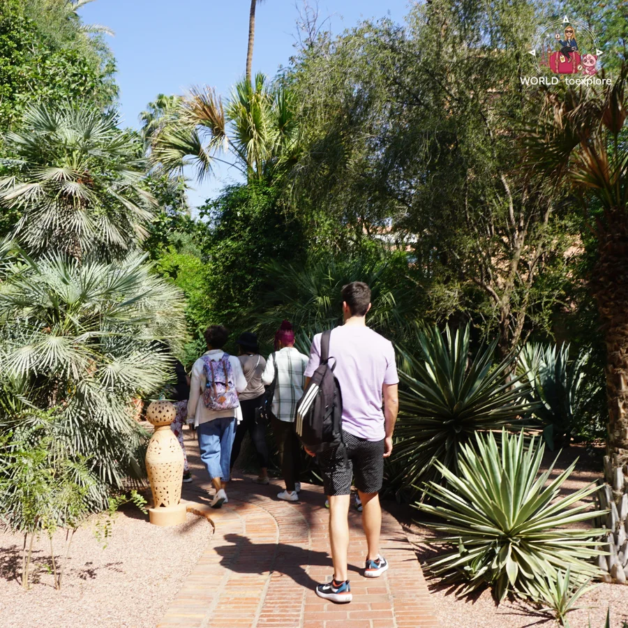 Visitors seen from behind walking along a terracotta path, among palm trees and agaves, in the garden