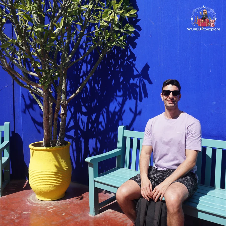 Man sitting on a turquoise bench in front of an intense blue wall and a yellow pot with a tree