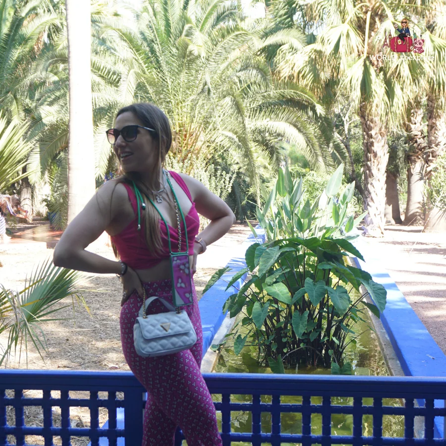 Woman in a pink top and patterned trousers, hand on her hip, next to a narrow pool and a blue railing, among palm trees