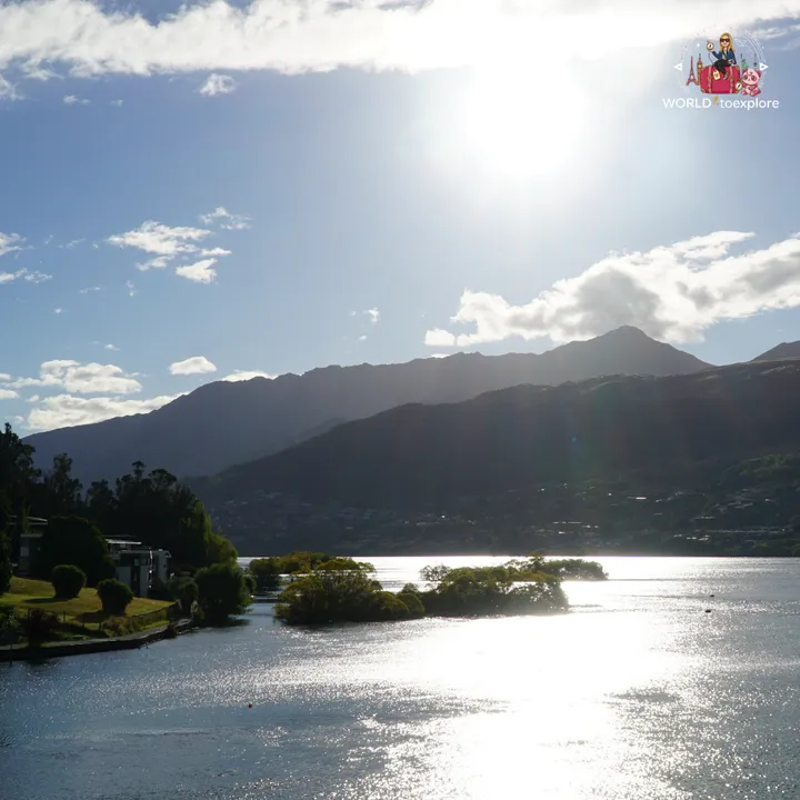 View of Lake Wakatipu against the sun, with small tree-covered islets and the Queenstown mountains in the background