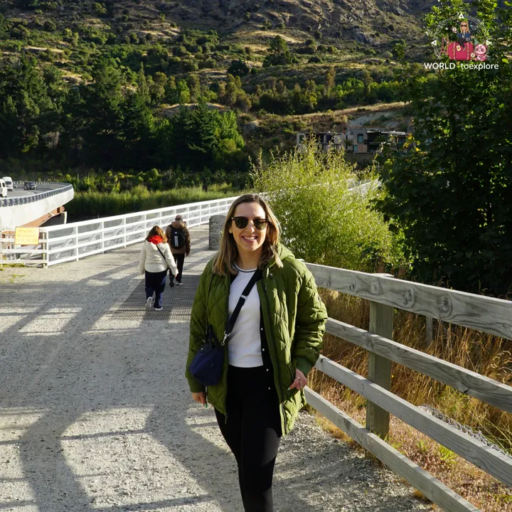 The author walking across the wooden bridge at the Kawarau Falls Dam, with mountains and trees in the background