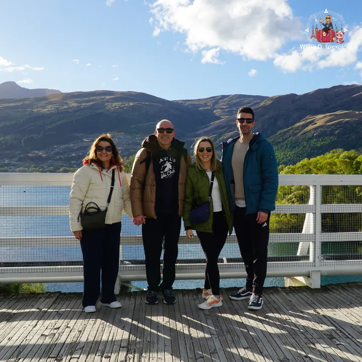 A group of four people holding hands on the Kawarau Falls Dam bridge, with the lake and Queenstown mountains in the background