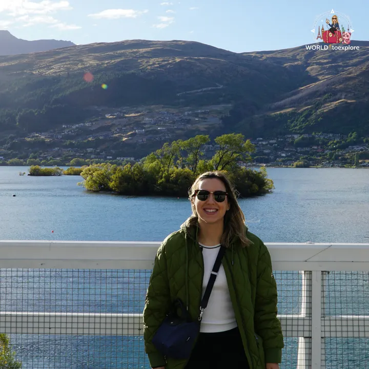 The author smiling on the Kawarau Falls Dam bridge, with a small tree-covered islet and Lake Wakatipu in the background