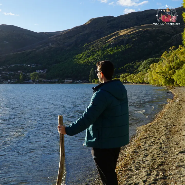 A man in a teal jacket, leaning on a wooden stick, looking out over Lake Wakatipu from the beach