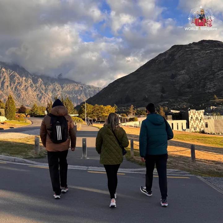 A group walking away from the camera along a lakeside path, with the Queenstown mountains in the background