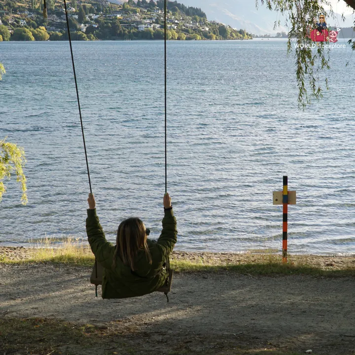 The author on a swing by Lake Wakatipu, facing the water and the houses on the opposite shore