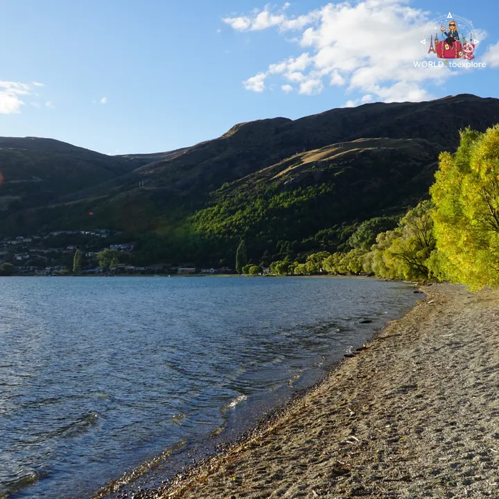 Sandy shore of Frankton Beach by Lake Wakatipu, with trees and mountains in the background