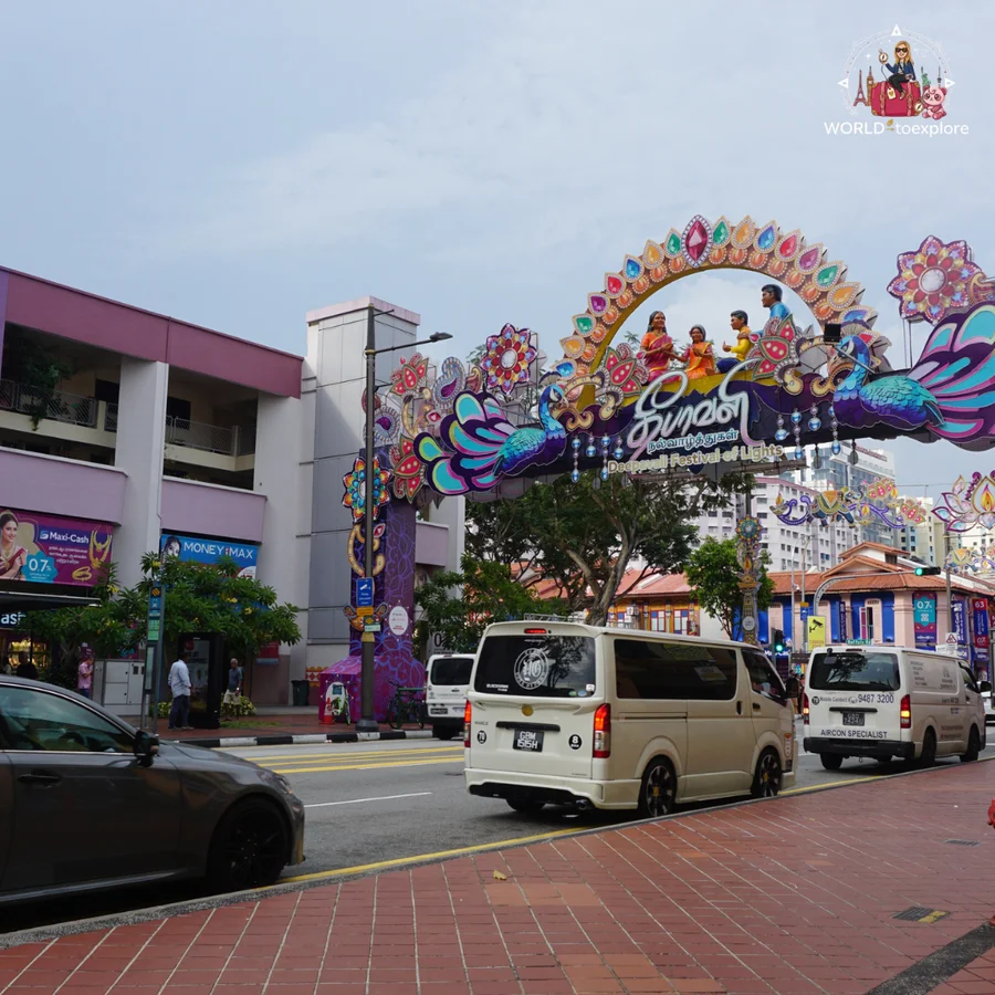 A colourful decorative arch, with peacocks and figures, over a street in Little India, Singapore, with vans driving past