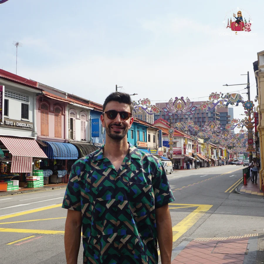 Her husband in a patterned shirt and sunglasses on a street in Little India, with colourful decorations over the road and old shops behind him