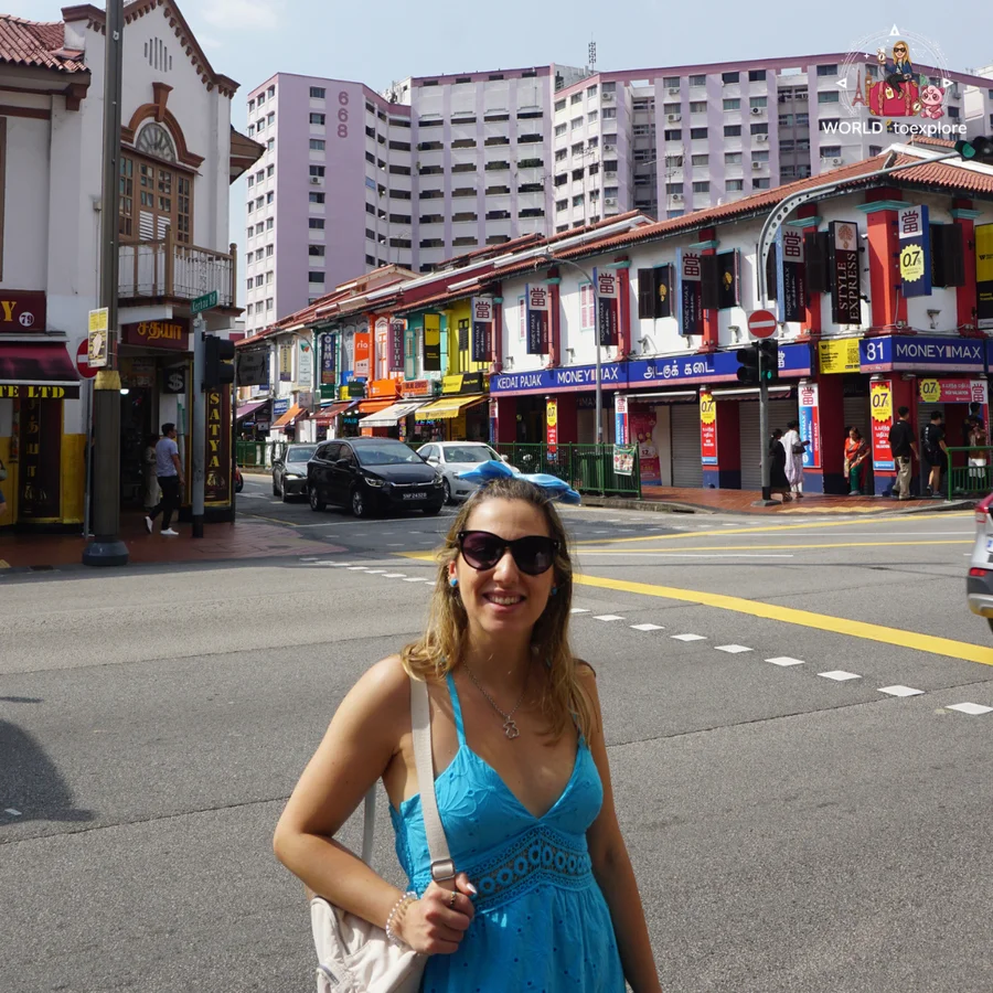 Cristina smiling, in a blue dress, at a junction in Little India, with colourful shopfronts and tall buildings behind her