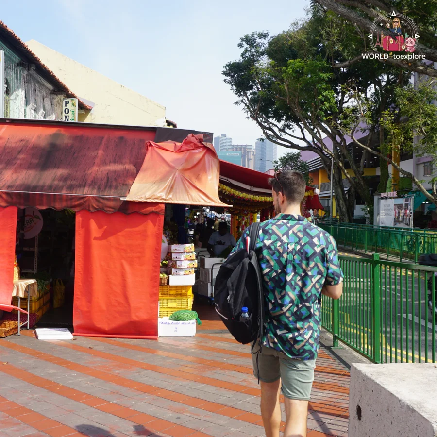 Her husband with a backpack walking past a stall with a red awning on a street in Little India