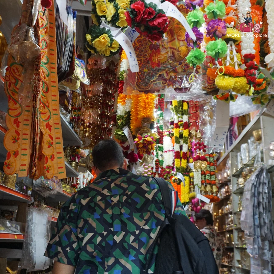 Her husband seen from behind inside a shop in Little India full of flower garlands, fabrics and colourful hanging decorations