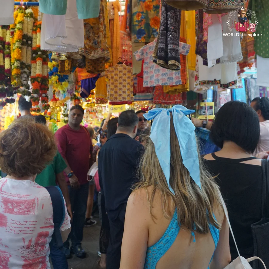 Cristina seen from behind, with a blue bow in her hair, in the middle of a crowd on a shopping street in Little India, with fabrics and garlands hanging above