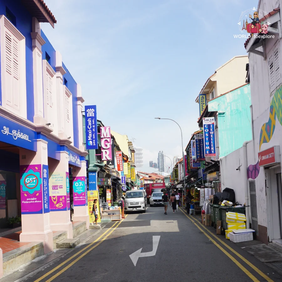 A narrow street in Little India with blue and white shopfronts, colourful signs and Singapore's skyscrapers in the background