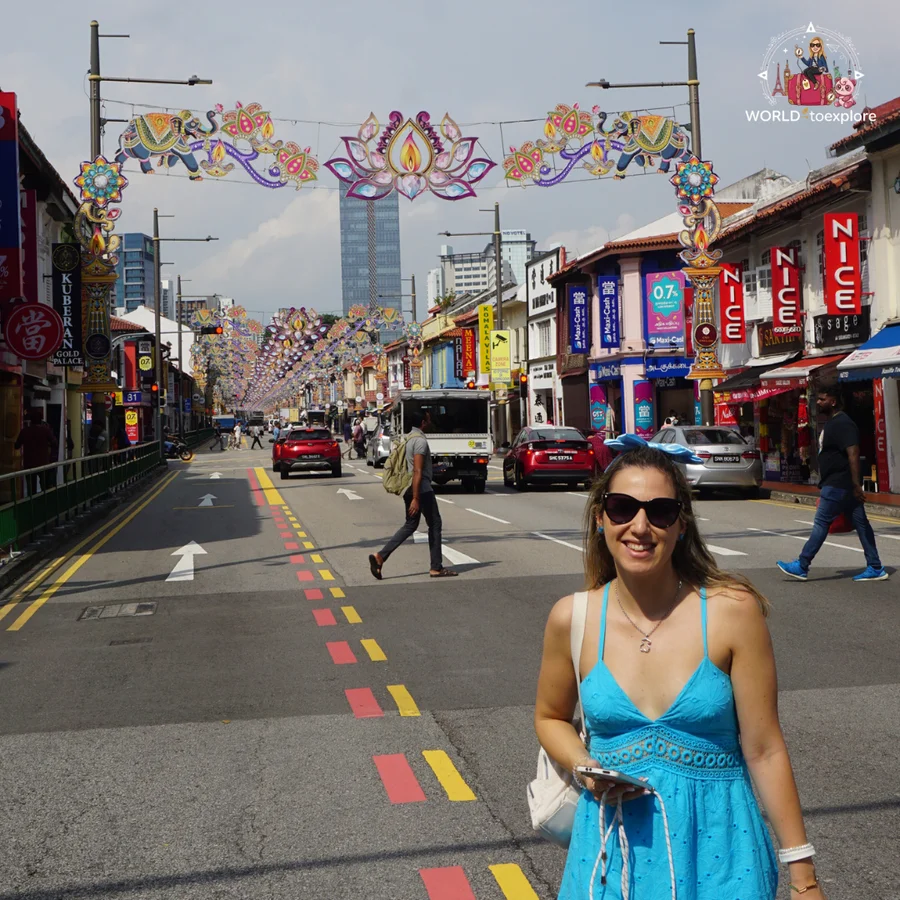 Cristina in a blue dress and sunglasses on a street in Little India, with decorative lotus-flower arches and colourful shops behind her
