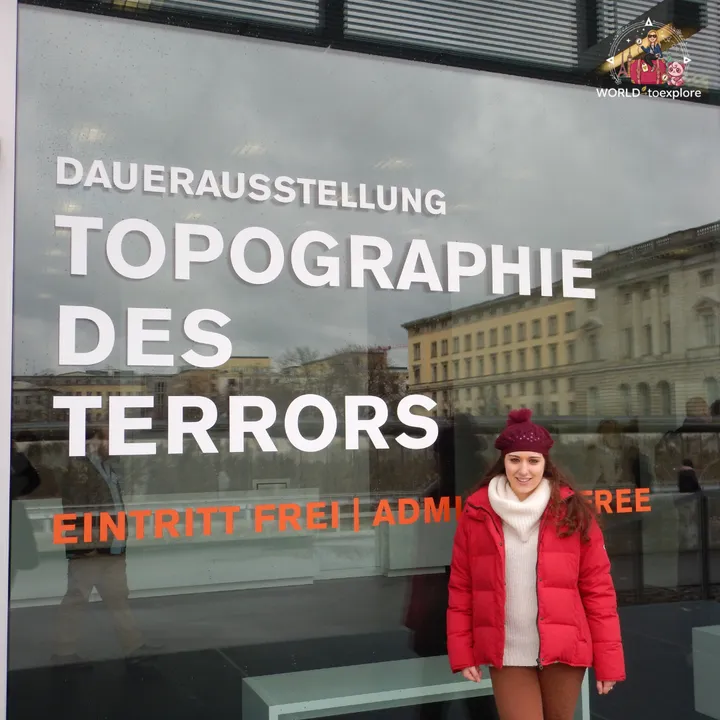 In front of the glass facade of the Topography of Terror, with the name of the permanent exhibition and the free admission notice