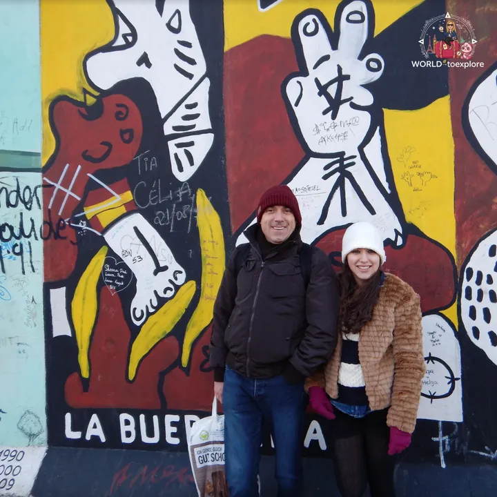 Two people wrapped up in coats and hats in front of an East Side Gallery mural painted in red, yellow and white