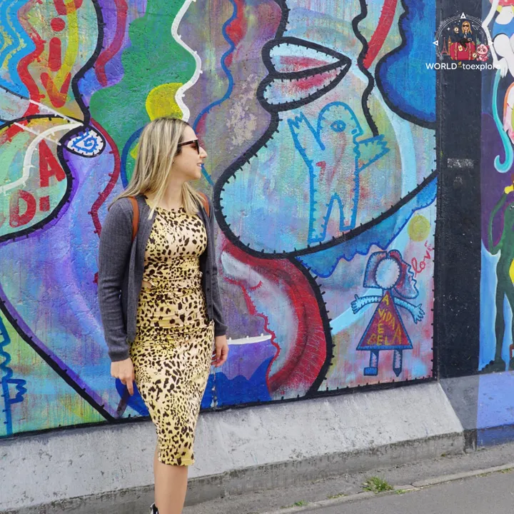 Standing in front of a brightly coloured East Side Gallery mural, with thick-outlined figures and the words A VIDA É BELA painted on one of them