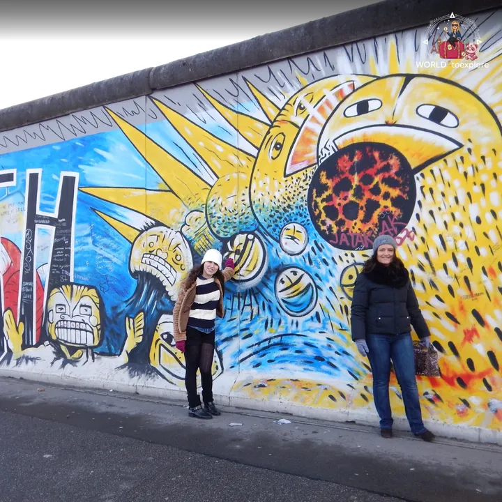 Two people in winter coats in front of a yellow and blue East Side Gallery mural, with screaming painted faces and a large yellow figure