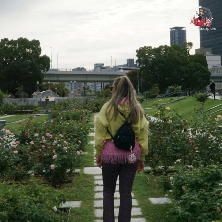 Woman walking away along a path in the Nakanoshima Rose Garden, with a bridge and the Osaka skyline behind