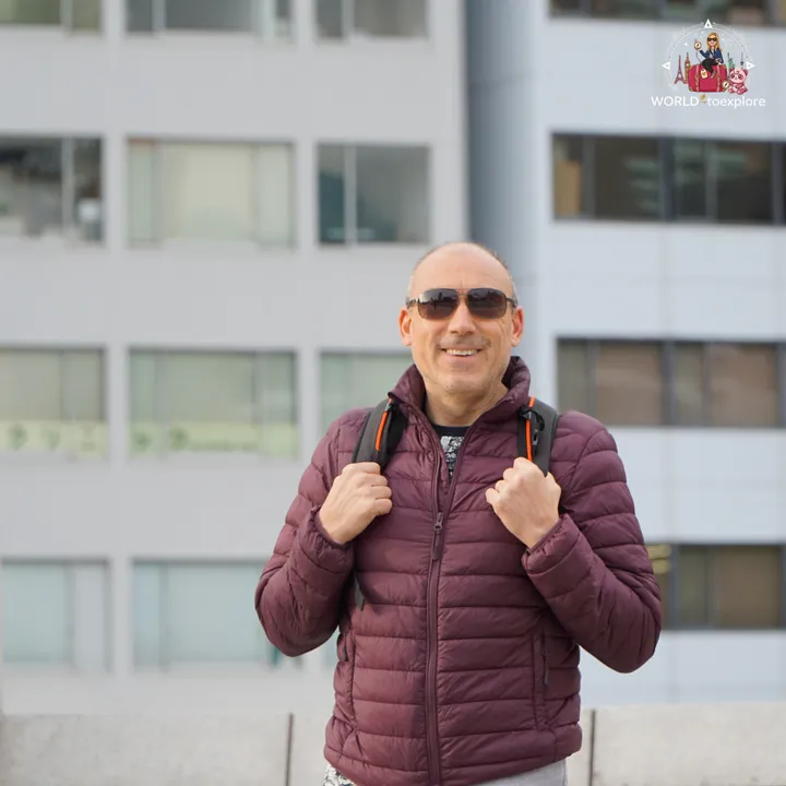 Smiling man with a backpack, standing in front of a modern glass building in Osaka