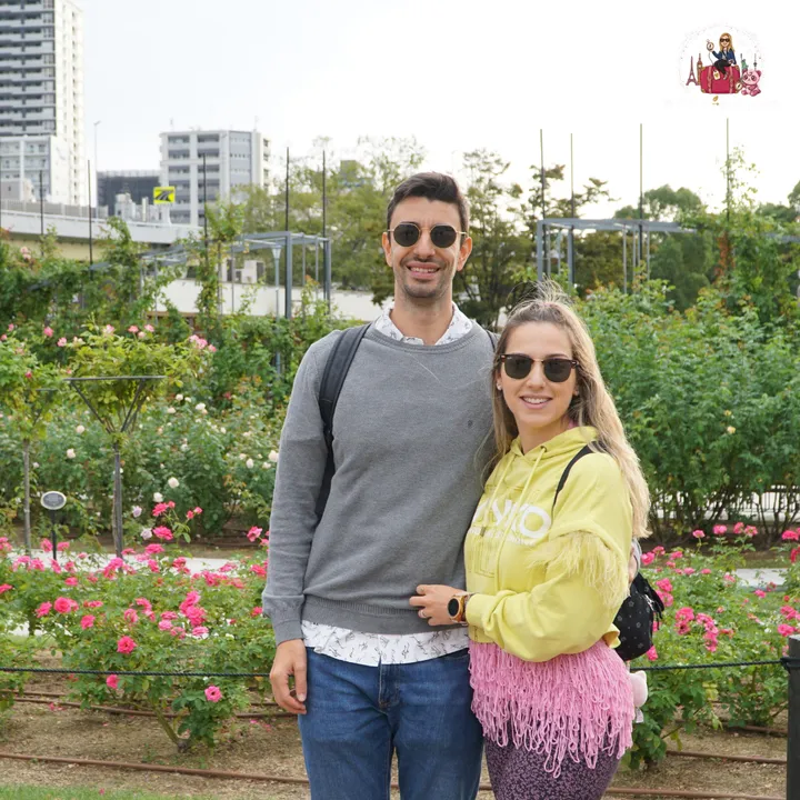 Smiling couple at the Nakanoshima Rose Garden, surrounded by rose bushes, with the Osaka skyline behind